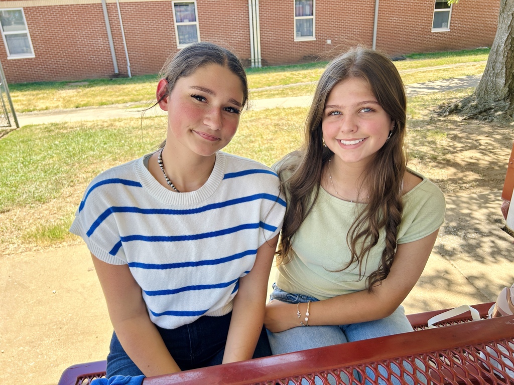 Two girls sitting on a bench outside, one wearing a white and blue shirt, the other a yellow top. Behind them is a brick building with windows and grass.