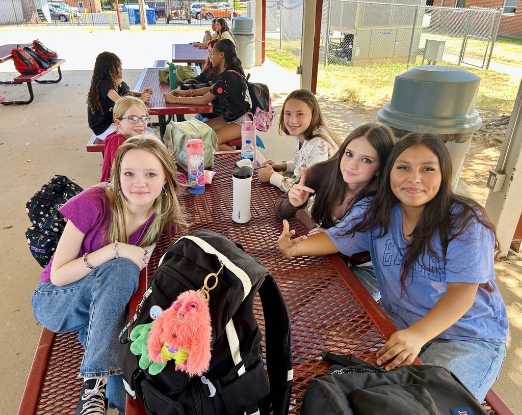 A group of girls are sitting on a bench with backpacks in an outdoor eating area.