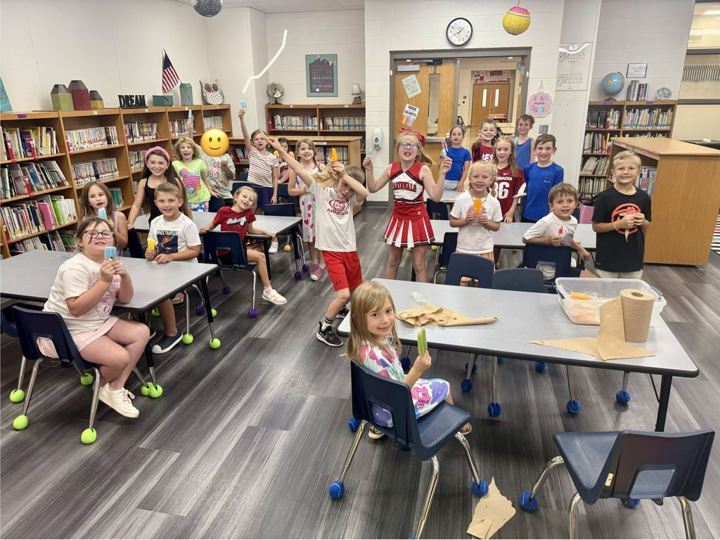 A classroom with tables and chairs, children seated and standing. They are in a school library with shelves of books and a flag.