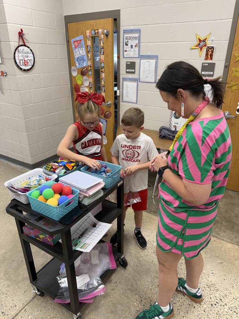 A teacher assists two children at a cart with various craft supplies. A bulletin board is in the background.