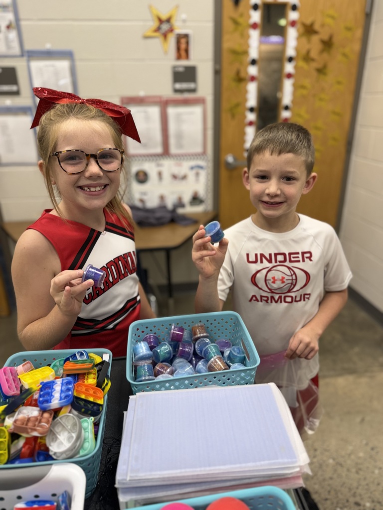 Two children in a classroom holding items. A girl in a red cheerleading uniform and a boy in a white Under Armour shirt.
