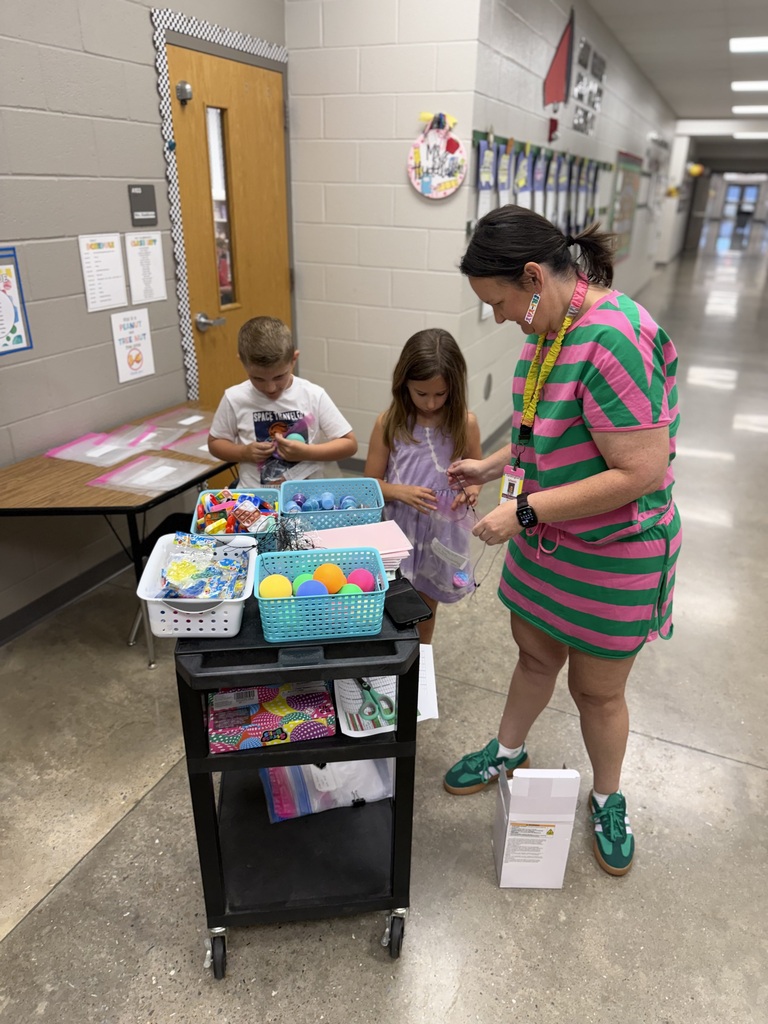 A woman helps two children with crafts in a school hallway with a table and various items.