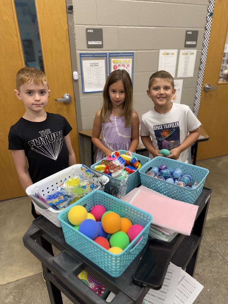 Three children stand in a classroom, next to a table with bins full of colorful balls and items.
