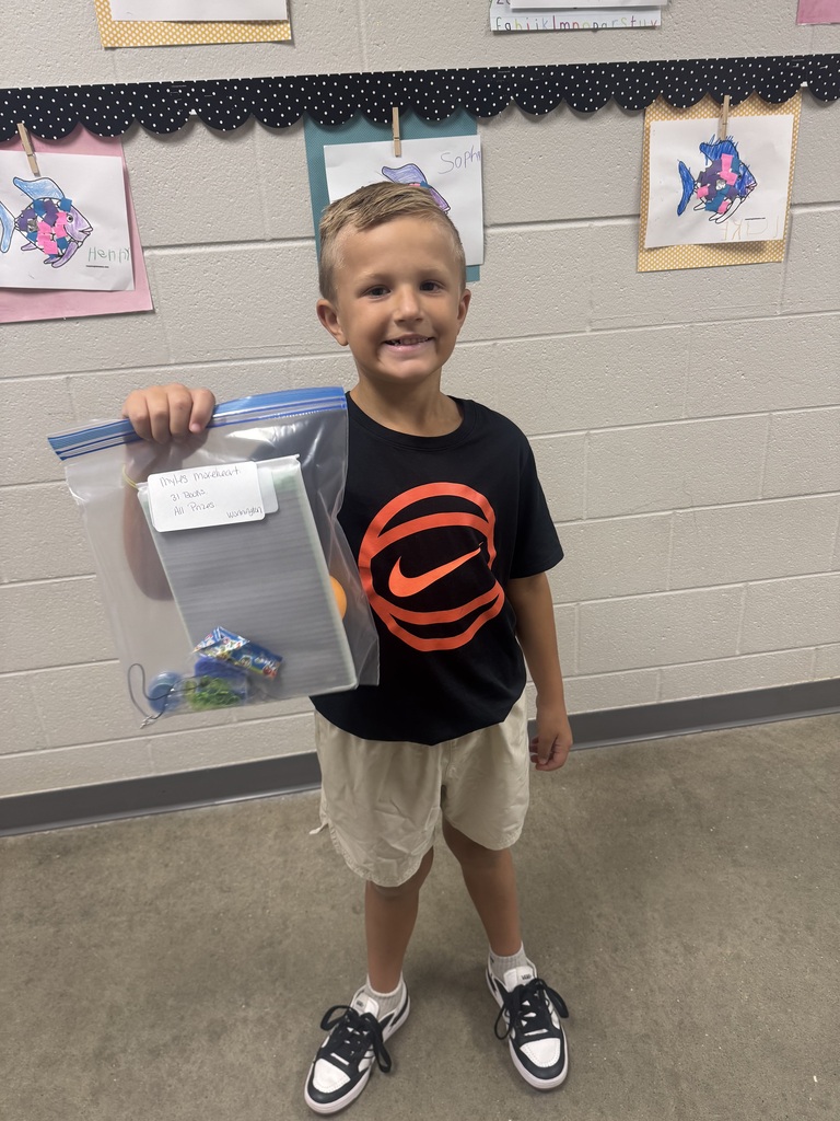 A young boy in a black shirt and shorts holds a plastic bag containing a small object. He stands in a room with a white wall and children's artwork.