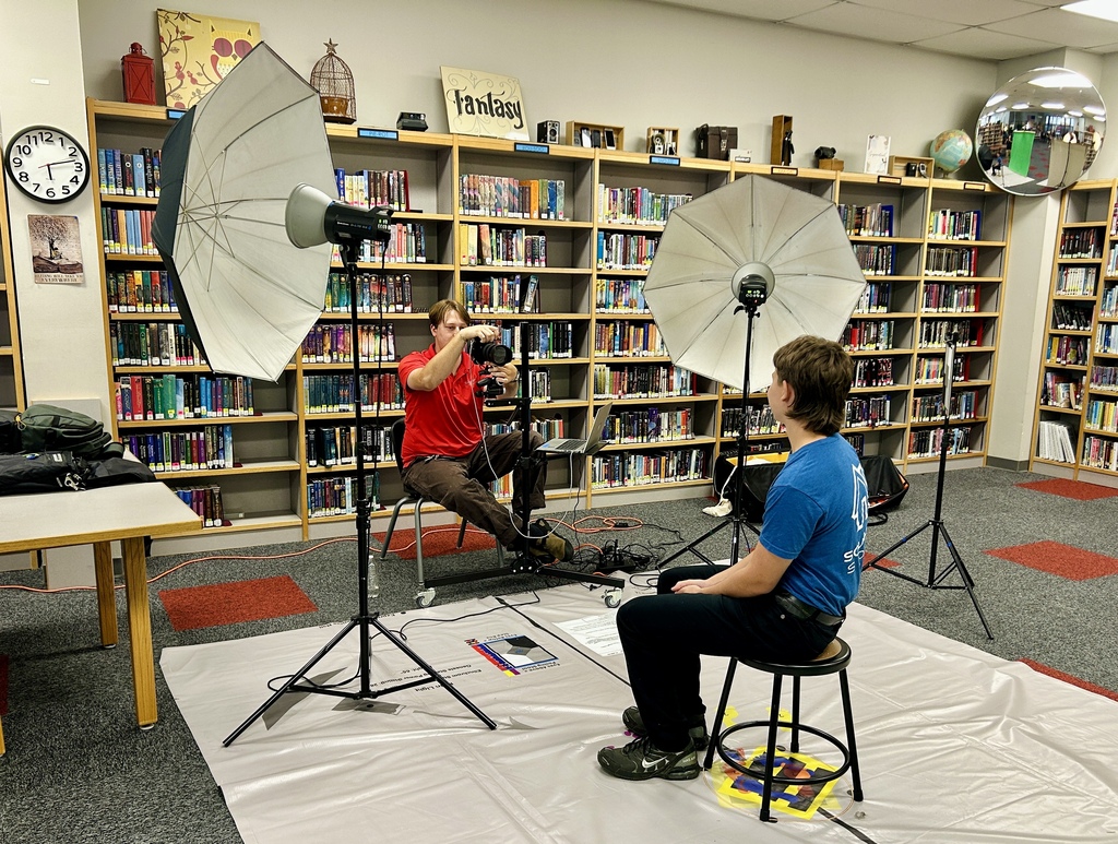 Two people in a library. One is sitting on a stool, the other is on a chair, both in front of camera equipment.