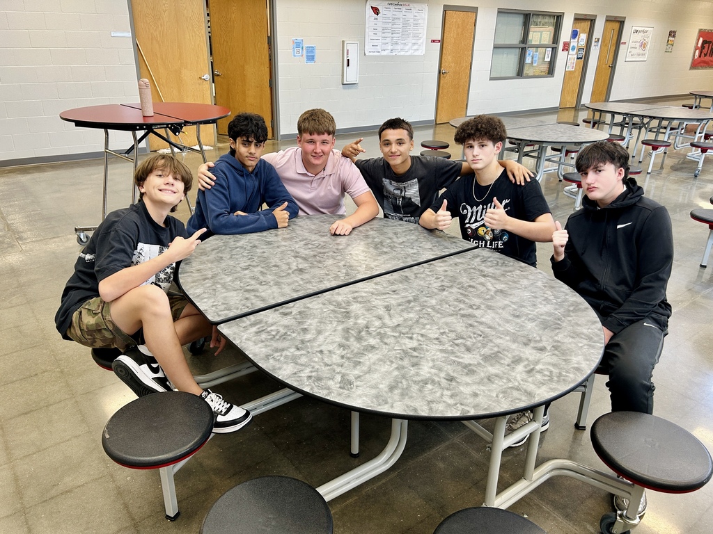 Six teens pose for a photo at a rectangular table in a school cafeteria. The table has two benches on each side.