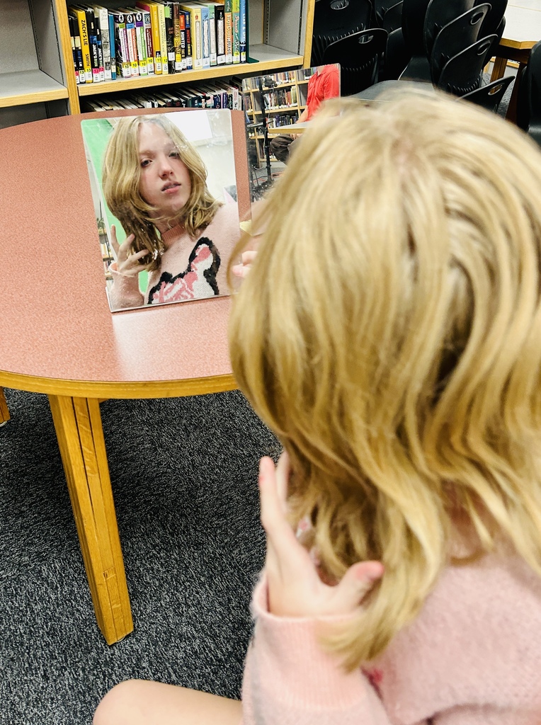 A child with blonde hair examines their reflection in a mirror, situated in a room with bookshelves and chairs.