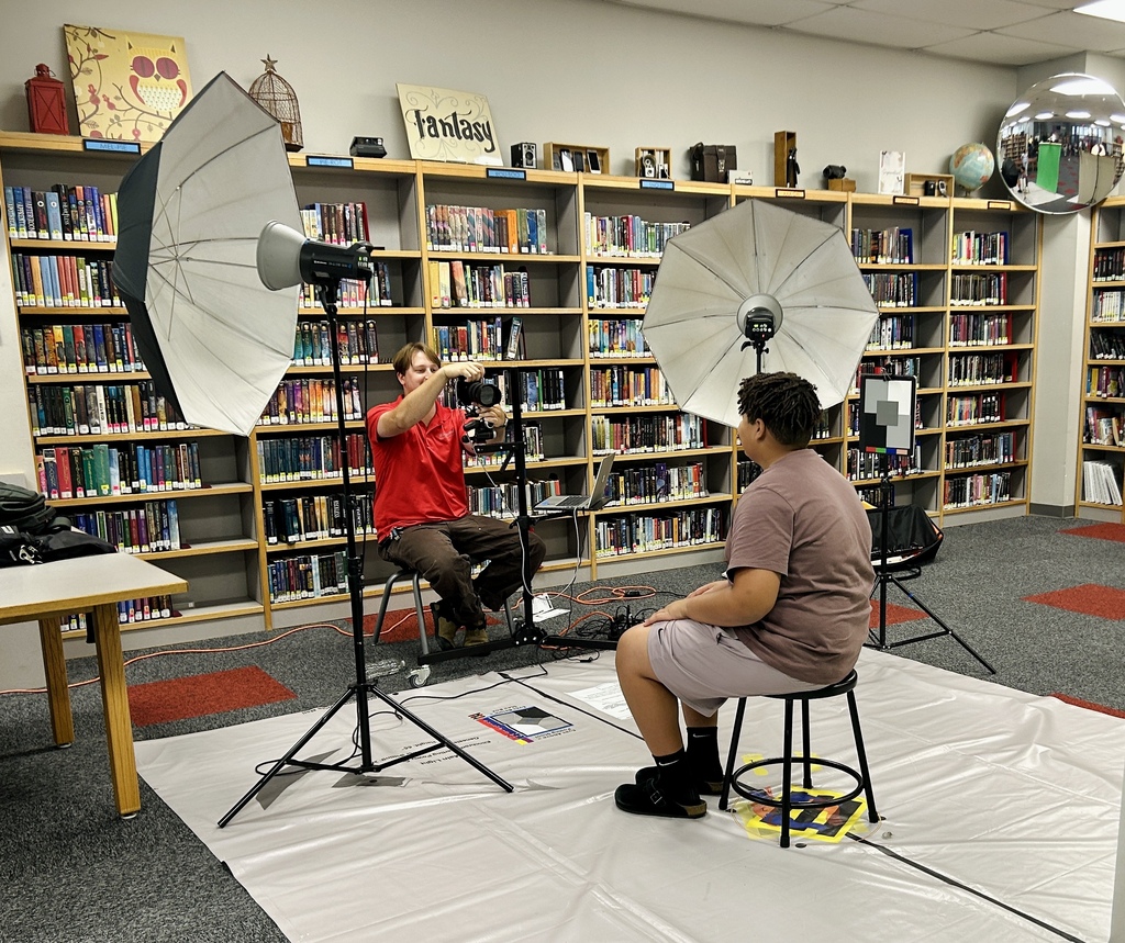 Two people in a library. One is seated with a camera, and the other is on a stool. Photo equipment is set up on a white sheet.