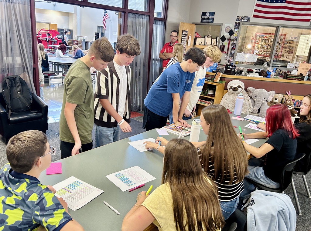 Several students gather around a table in a classroom, working on papers. Background includes shelves and an American flag.
