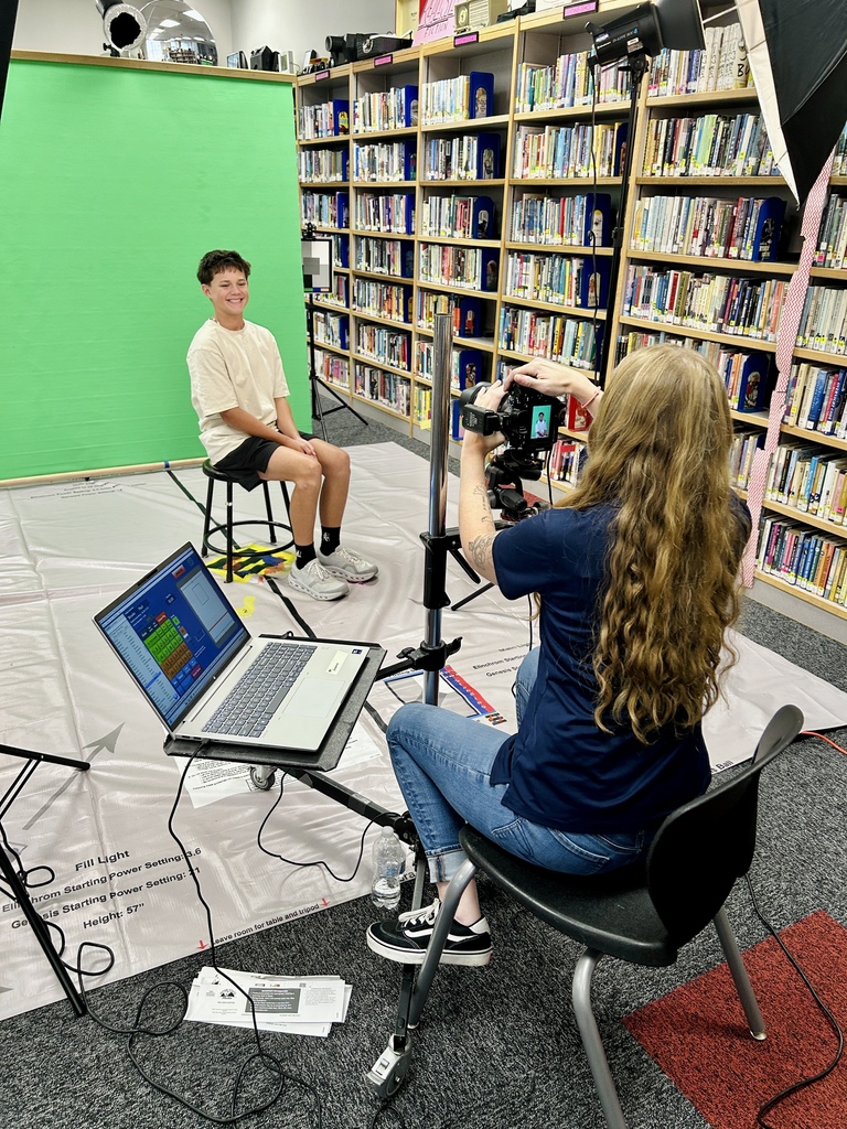 In a library, a woman films a seated young man using a camera on a tripod. A laptop sits on a desk.