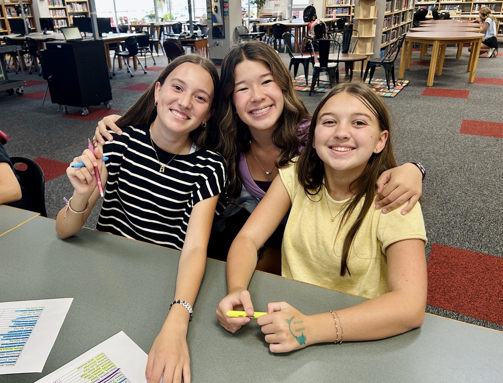 Three girls in a library, two smiling and leaning on a table, the third with a yellow pen.
