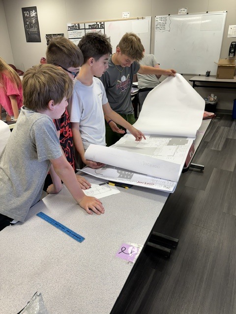 Five children examine a large sheet of paper on a table, with a whiteboard in the background.