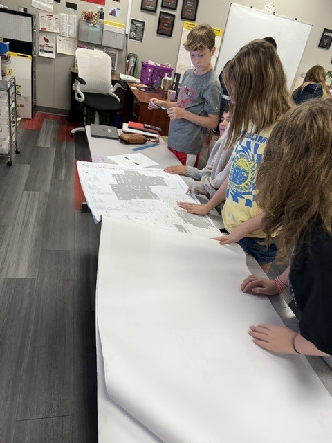 A group of students examines a large piece of paper on a table. Background contains shelves, a desk, and a chair.