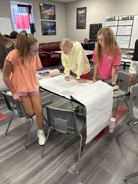Three girls work on a project at a table in a room with gray chairs, gray carpet, and a whiteboard.