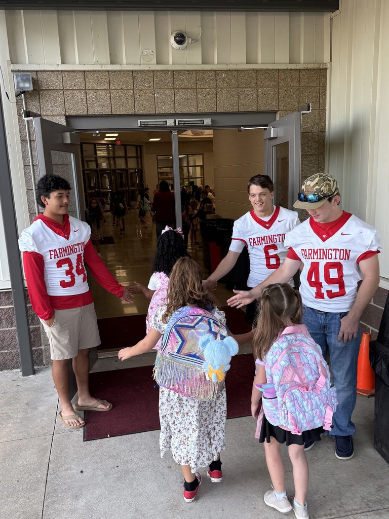 Five people, including two boys in sports jerseys, assist young children with their backpacks at a school entrance.