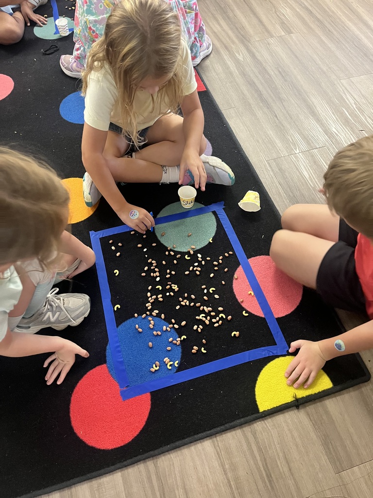 Three children are playing on a rug with a blue square and colored circles. One child holds a cup.