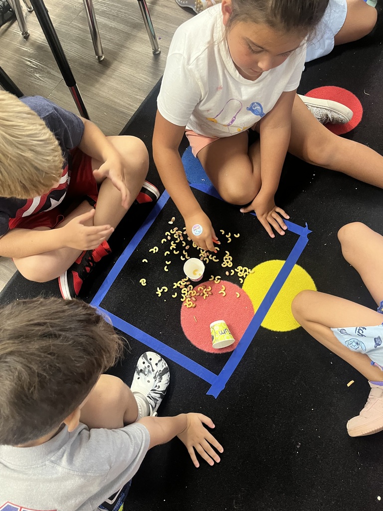 Children seated on a floor, engaged in an activity involving small objects. A square is marked on the floor in blue.