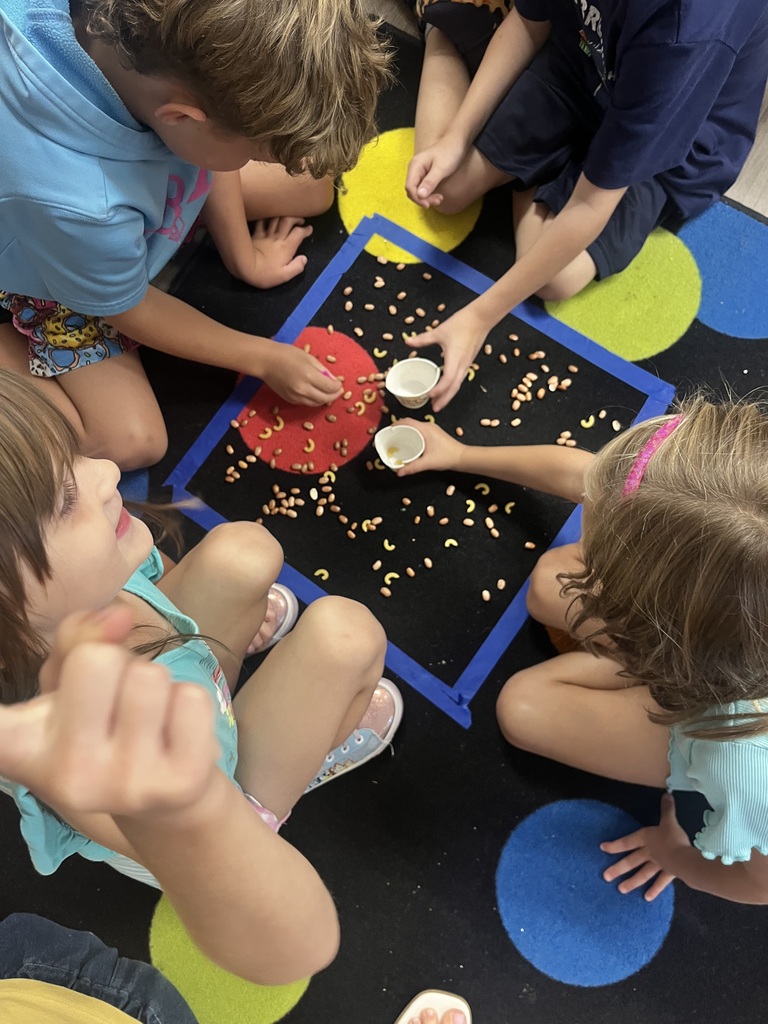 Several children sit on a colorful floor, handling small bowls with yellow objects, possibly seeds, on a carpet.