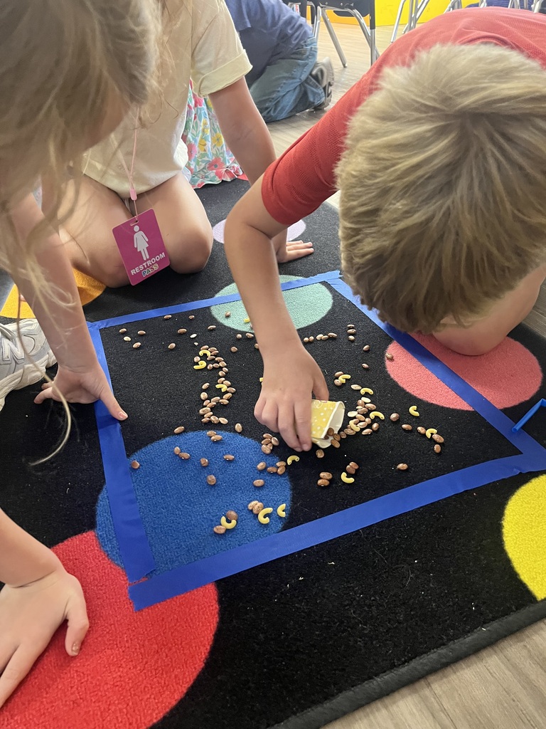 Children seated on the floor, one bent over a multicolored rug with circles and blue lines, picking up scattered cereal.