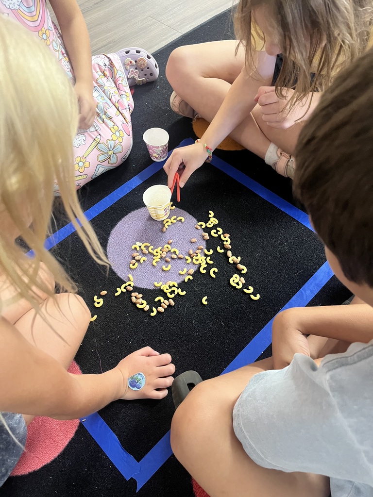 Three children sit on the floor and pick up yellow pasta to spell words. A blue outline is on the floor.