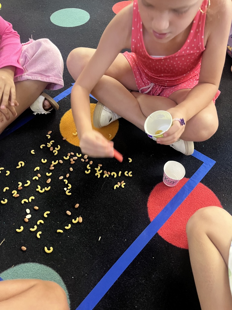 Child with red top and shorts plays with yellow shapes on floor, holds a cup, and sits with others.