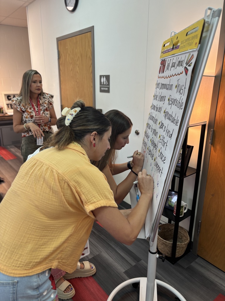 Two women writing on a white board with a colorful backdrop, one with a yellow shirt and another with a floral top.