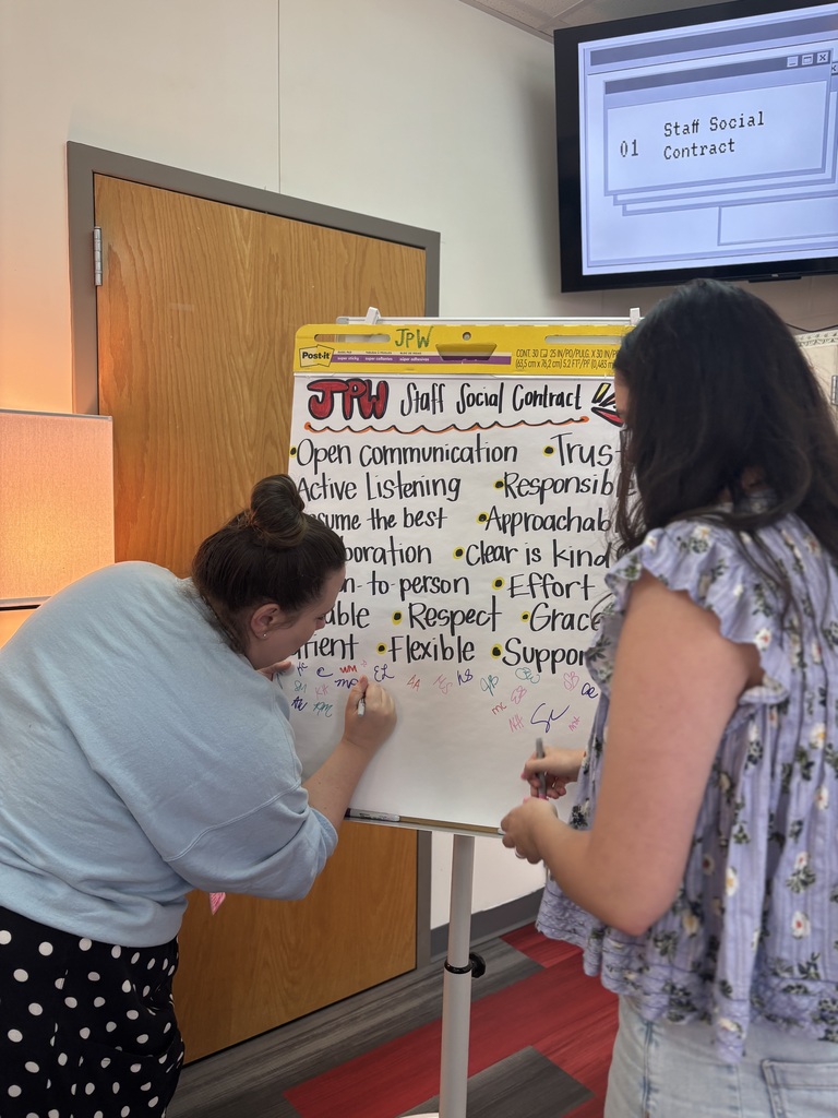 Two women in a room write on a whiteboard. The board has a list of words and phrases.