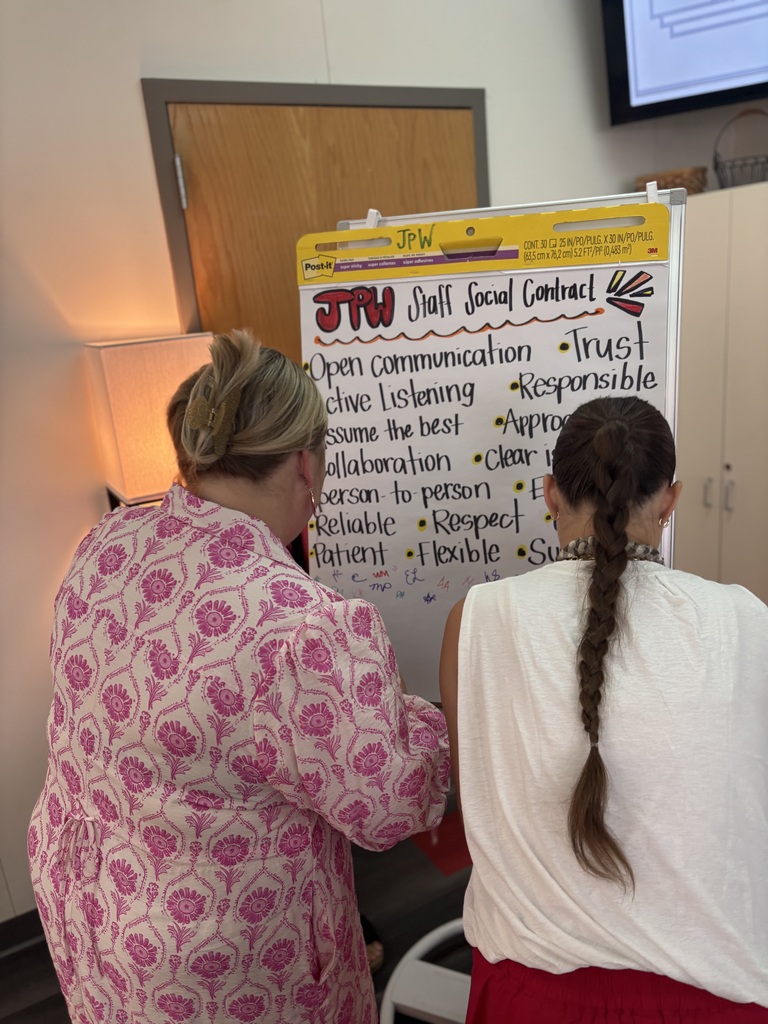 Two women stand before a whiteboard, discussing it. One wears a pink floral shirt. The other has long, braided hair.