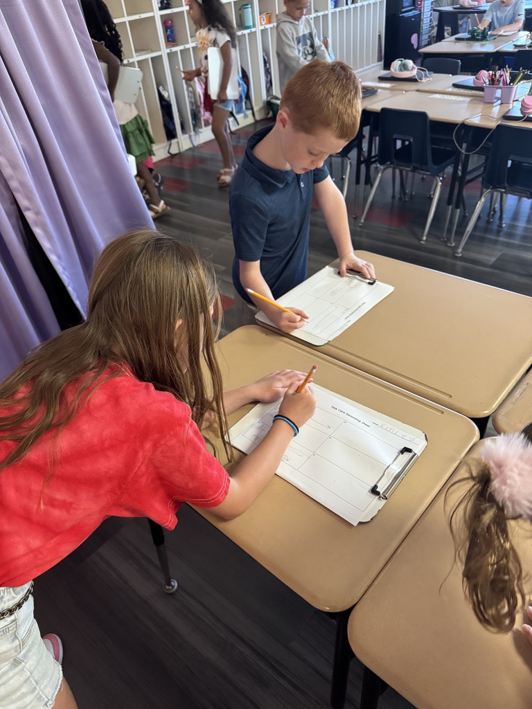 Two children work at desks, writing on paper, with a purple curtain and other desks in the background.