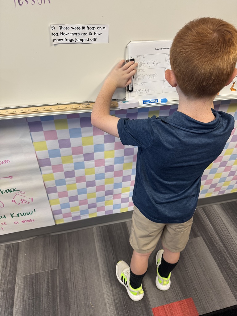 A child uses a marker to write on a whiteboard, standing in front of a colorful wall with a checkered pattern.