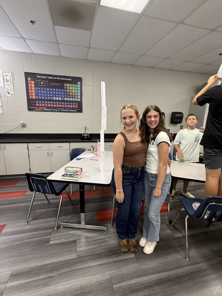 Two girls in a classroom. One girl has on a brown tank top and the other a white tee.