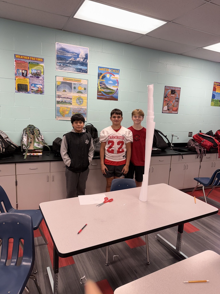 Three children in a classroom, two wearing football jerseys, standing around a table with a large tube.