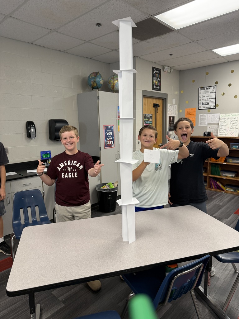 Three people stand by a table, holding items, with a tall tower of stacked paper cups in the center.
