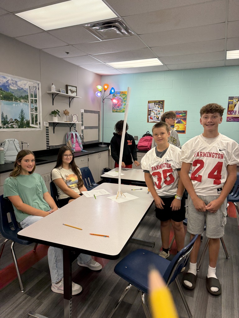 Several students in a classroom with light blue walls. Some sit around a table; others stand behind it.