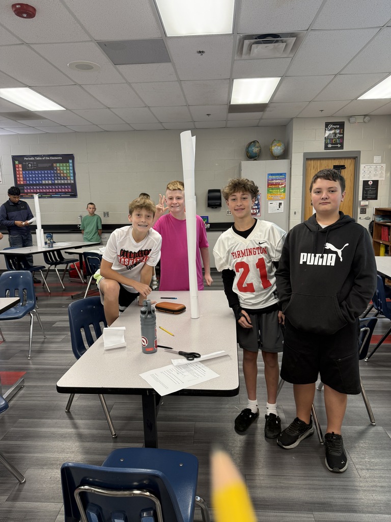 Four young boys stand around a table in a classroom, two holding a tall, thin object.