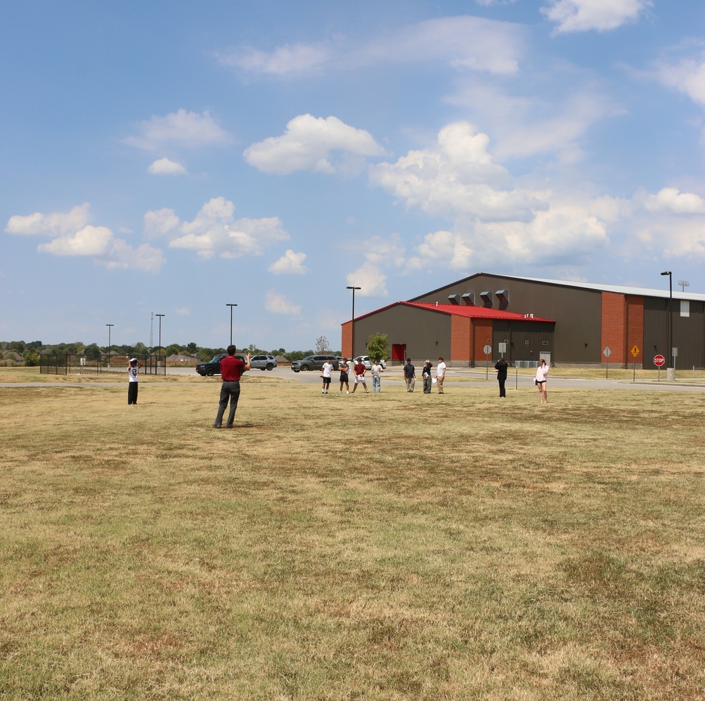 Field with several people. One in red shirt near center, others in groups. Large building in background with red trim.