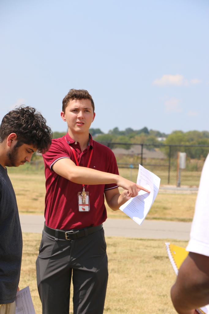 Two males in a grassy field. One holds a paper and points, the other looks at him. Trees and buildings are in the background.