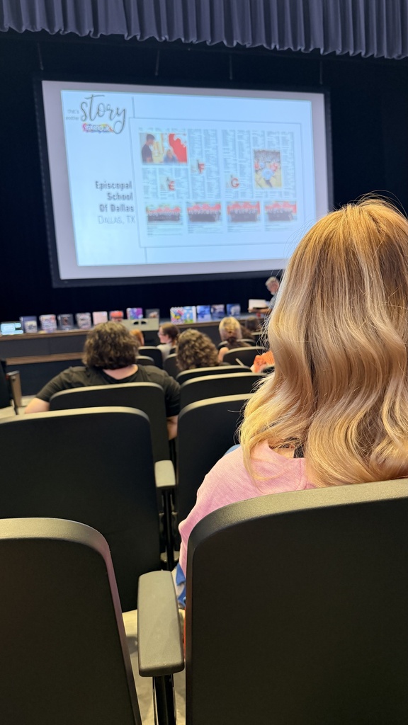 A seated audience watches a projector screen with text, people, and images. The chairs are black with armrests, and the background is blue.