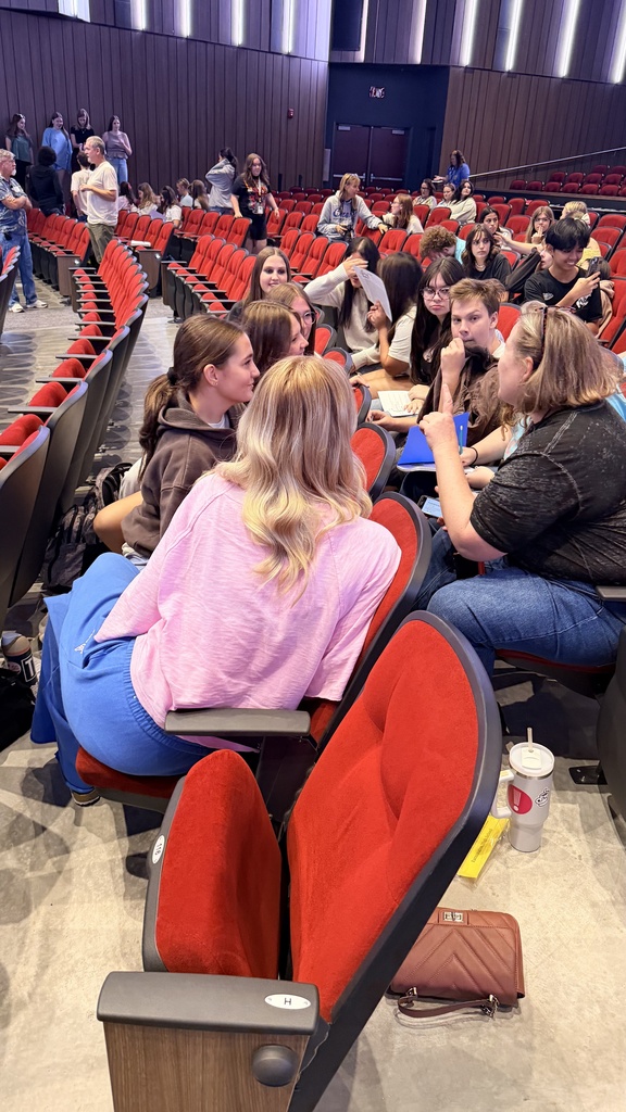 An auditorium with people sitting in rows of red chairs. A person in a pink shirt sits in the front row.