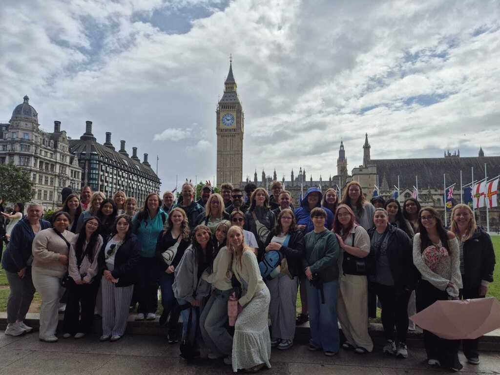 A group of people stands in front of a large clock tower on a cloudy day.