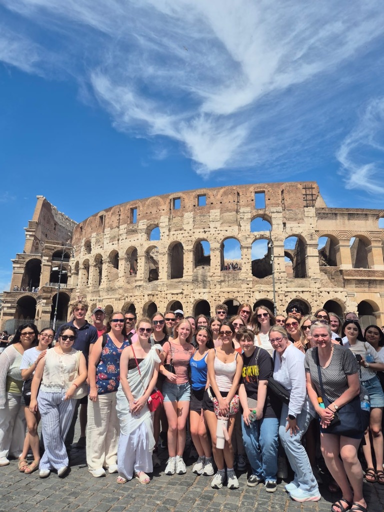 A group of people stands in front of the Colosseum in Rome under a blue sky.