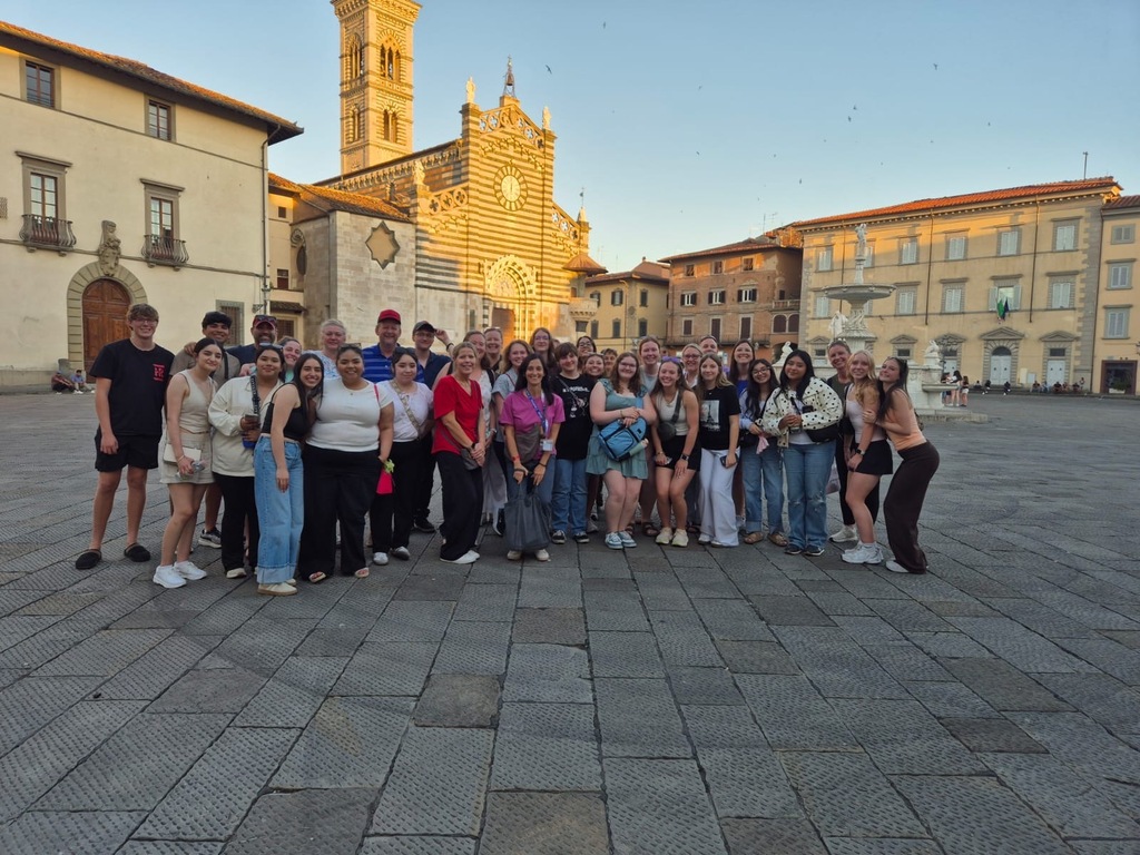 Group of people wearing various clothes, smiling and posing for a photo in a city square.
