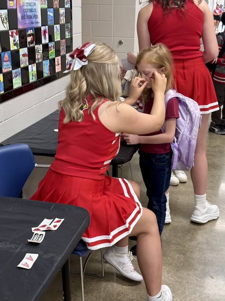 Woman in red cheerleading outfit sitting on chair applying makeup to girl in red uniform.
