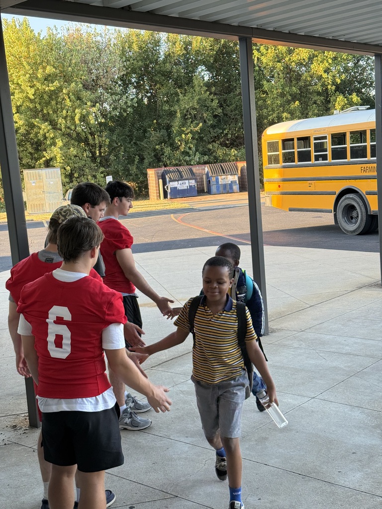 Students in numbered shirts greet a student with a backpack, near a school bus on the road.