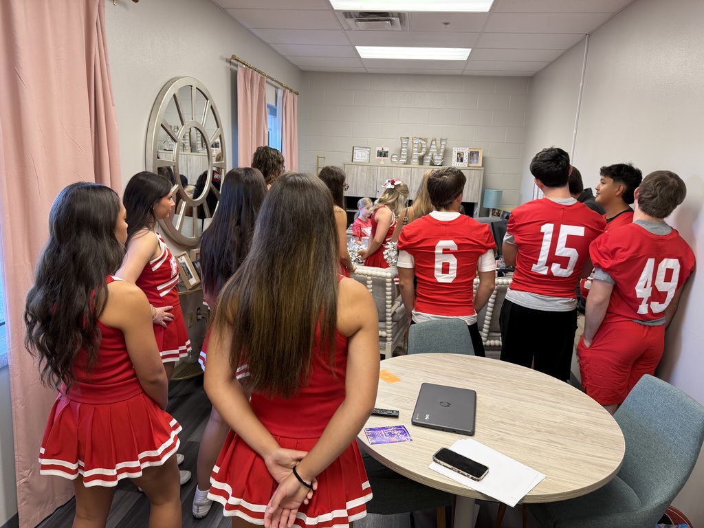 A group of people in red uniforms with numbers, standing around a table with a laptop.
