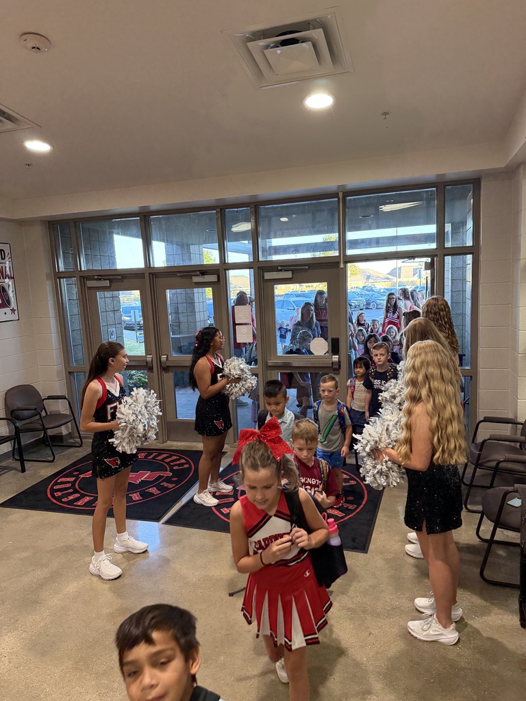 Several cheerleaders and children in a school hallway with large windows and chairs. Bright lights overhead.