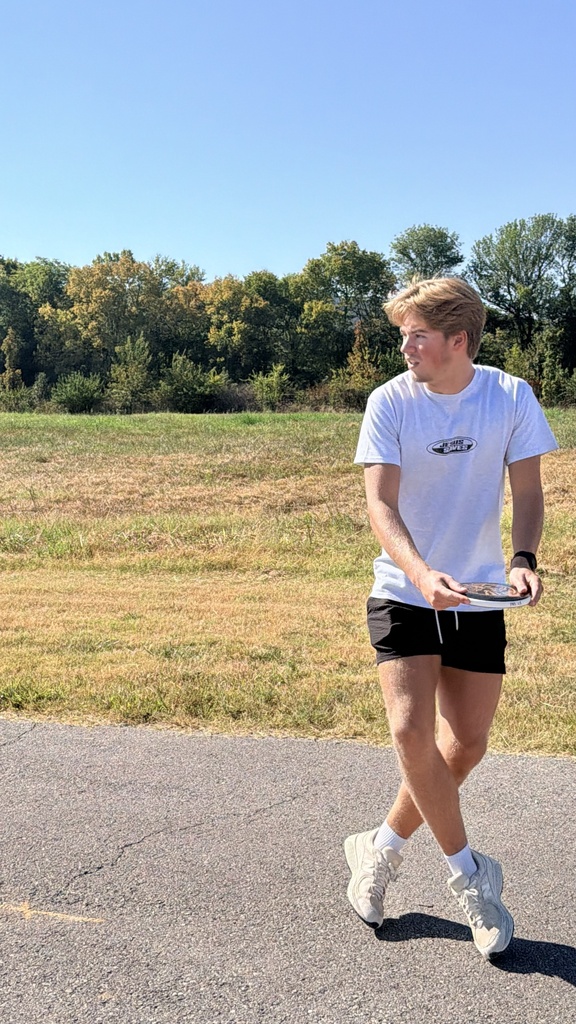 Man in a white shirt, black shorts, and white shoes holds a frisbee. Walking on a concrete path. Background has grassy field and trees.