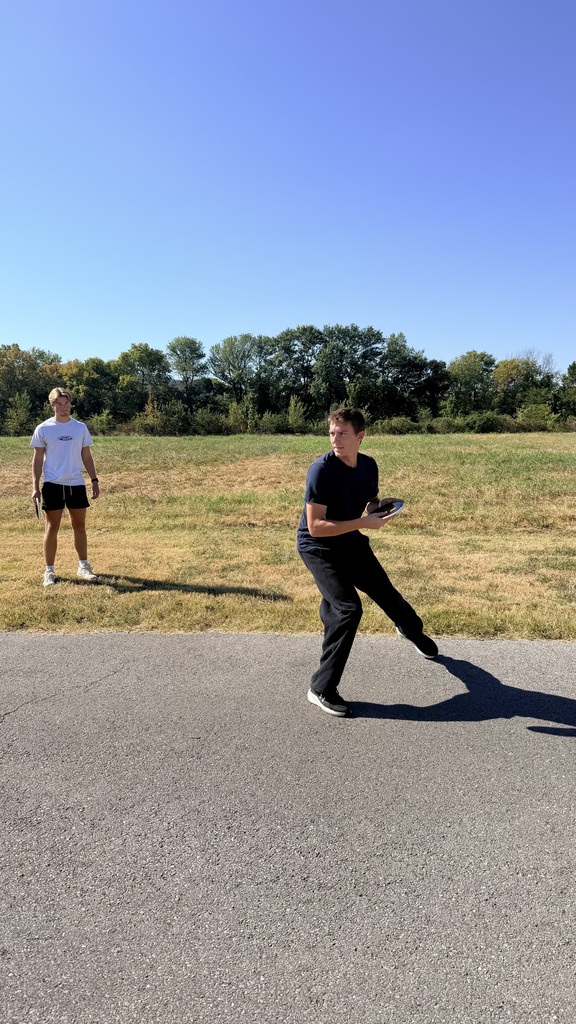 Two individuals in shorts and sneakers on a paved road with a grassy field and trees in the background.