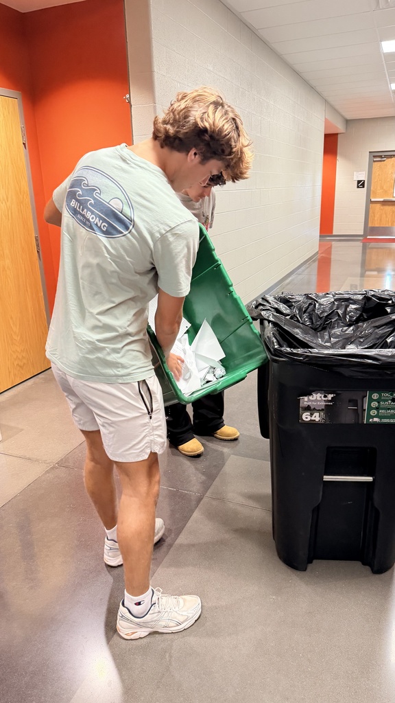 Person in white shorts and a t-shirt with a logo dumping a green bin into a trash can.