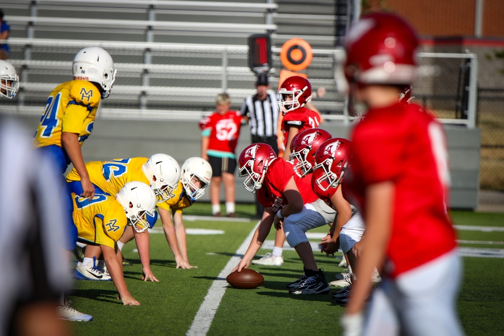 Youth football players, wearing red and yellow jerseys and helmets, line up on a field with a ball.